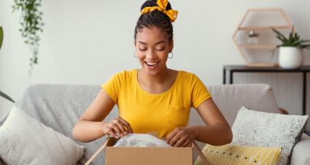 Quick Delivery Service Concept. Happy black lady received package, unpacking cardboard box, sitting on the sofa in living room at home, copy space.